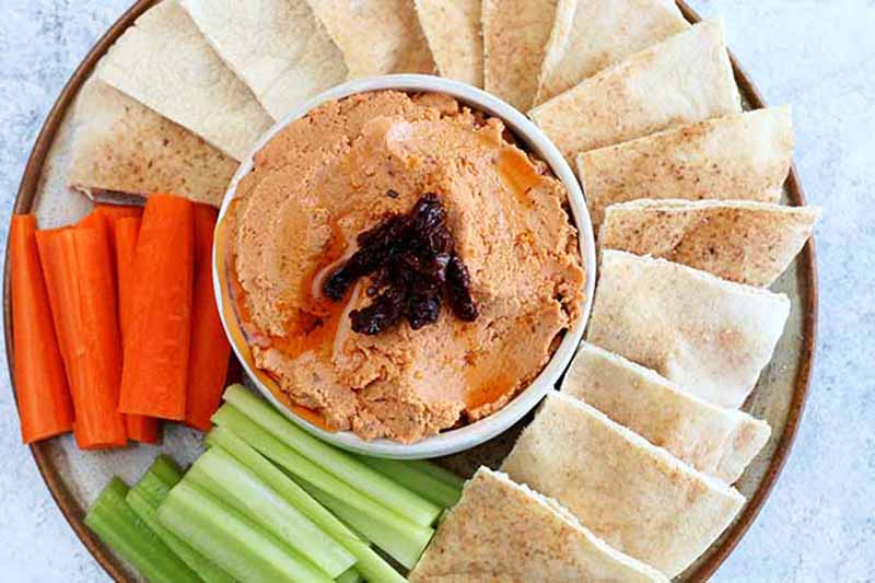 Horizontal top-down image of a white bowl filled with a light red pureed mixture on a plate with celery, carrots, and pita bread wedges.