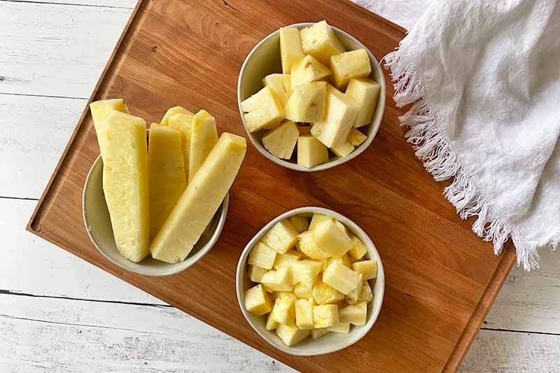 Horizontal image of different shapes of cut pieces of pineapple in white bowls on a wooden cutting board.