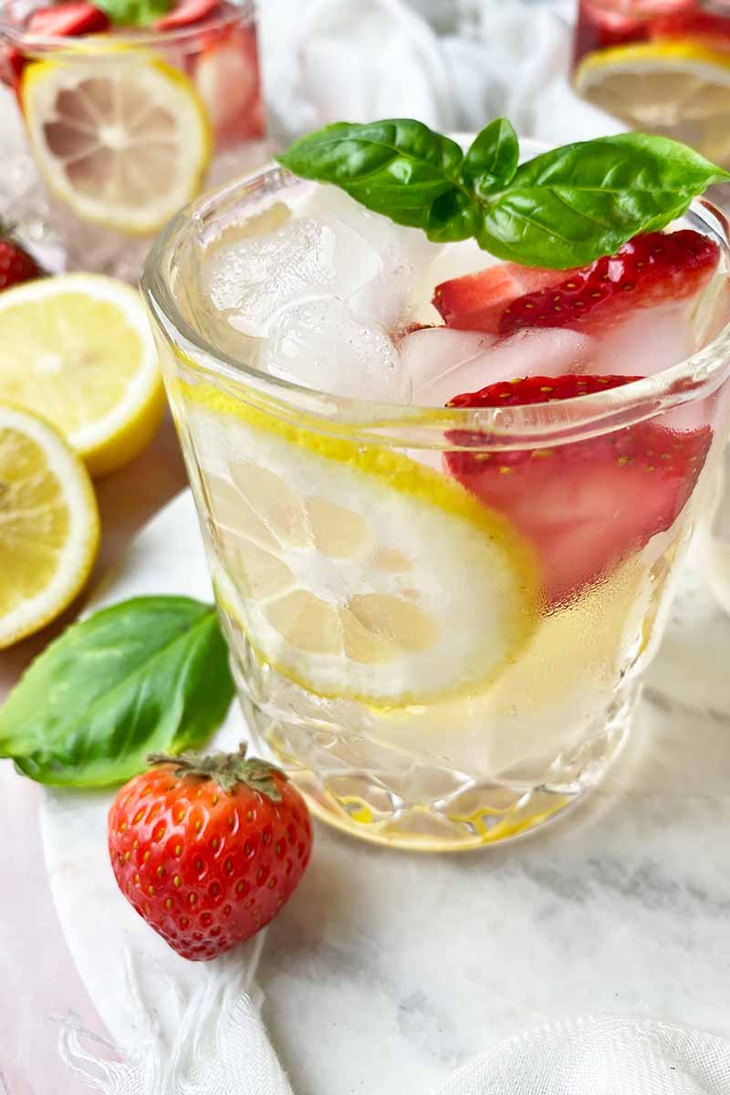 Vertical close-up image of a drink filled with a clear liquid mixed with lemon and strawberry pieces, with herb leaves as a garnish.