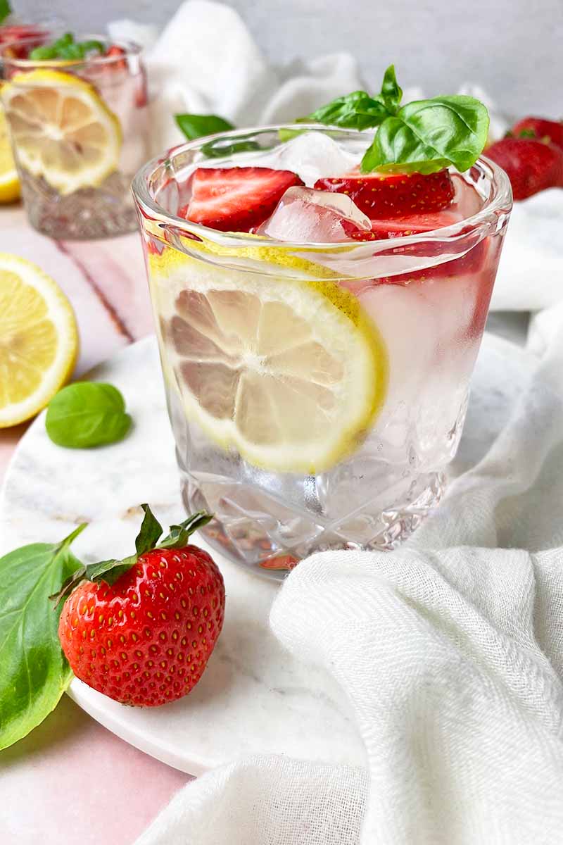 Vertical image of a glass filled with a clear beverage mixed with lemon and strawberries and garnished with herb leaves.
