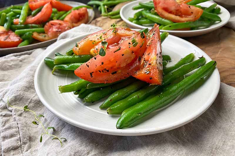 Horizontal image of a white plate with green beans, tomato wedges, and chopped thyme.