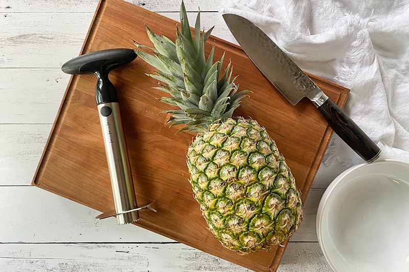 Horizontal image of a whole pineapple next to a coring tool and a knife on a board.