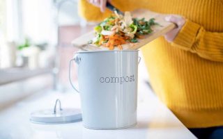 Horizontal image of a woman in a yellow sweater putting compost materials in a compost bin on a white countertop.