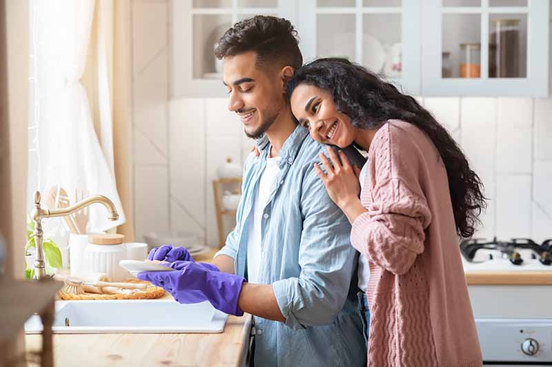 Horizontal image of a woman hugging a man while doing dishes.