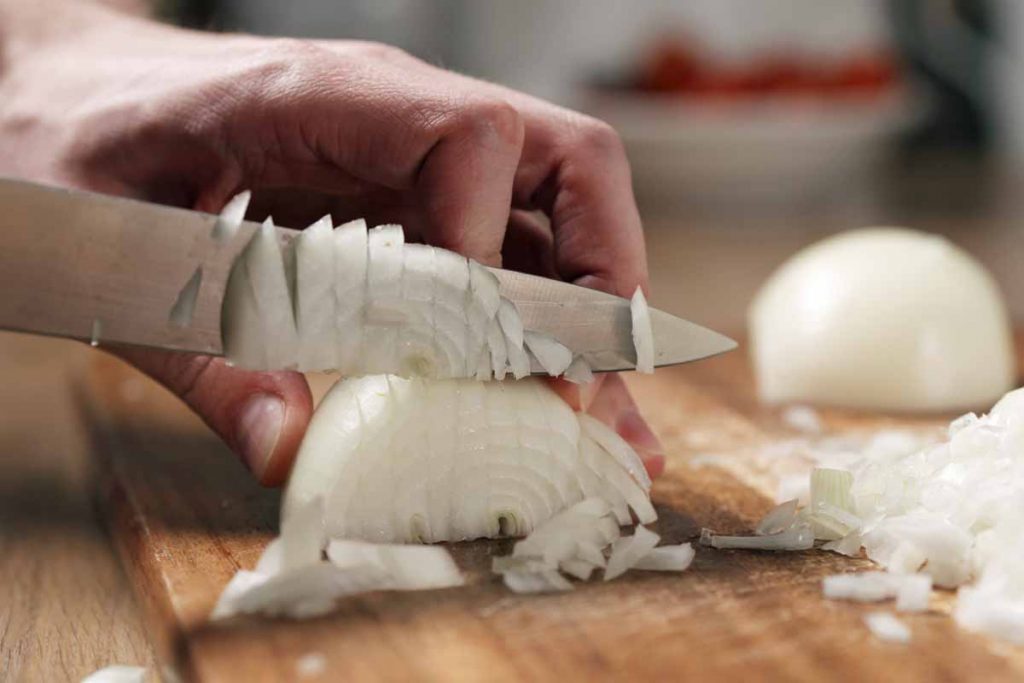 Man's hand cutting a white onion 
 bulb on a cutting board.