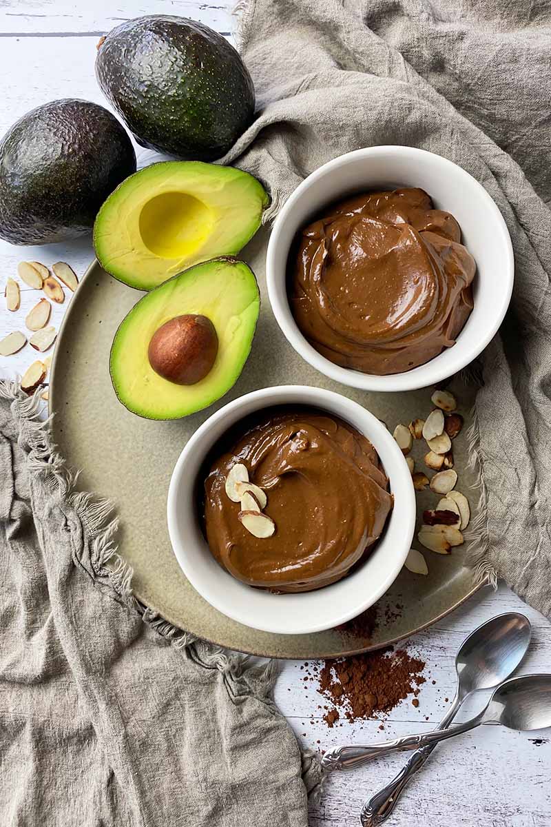 Vertical top-down image of two white bowls filled with a smooth and creamy chocolate dessert on a tan plate next to avocados.