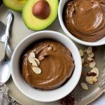 Horizontal top-down image of two white bowls filled with a creamy cocoa dessert, one topped with sliced almonds, on a plate next to a halved avocado and spoons.