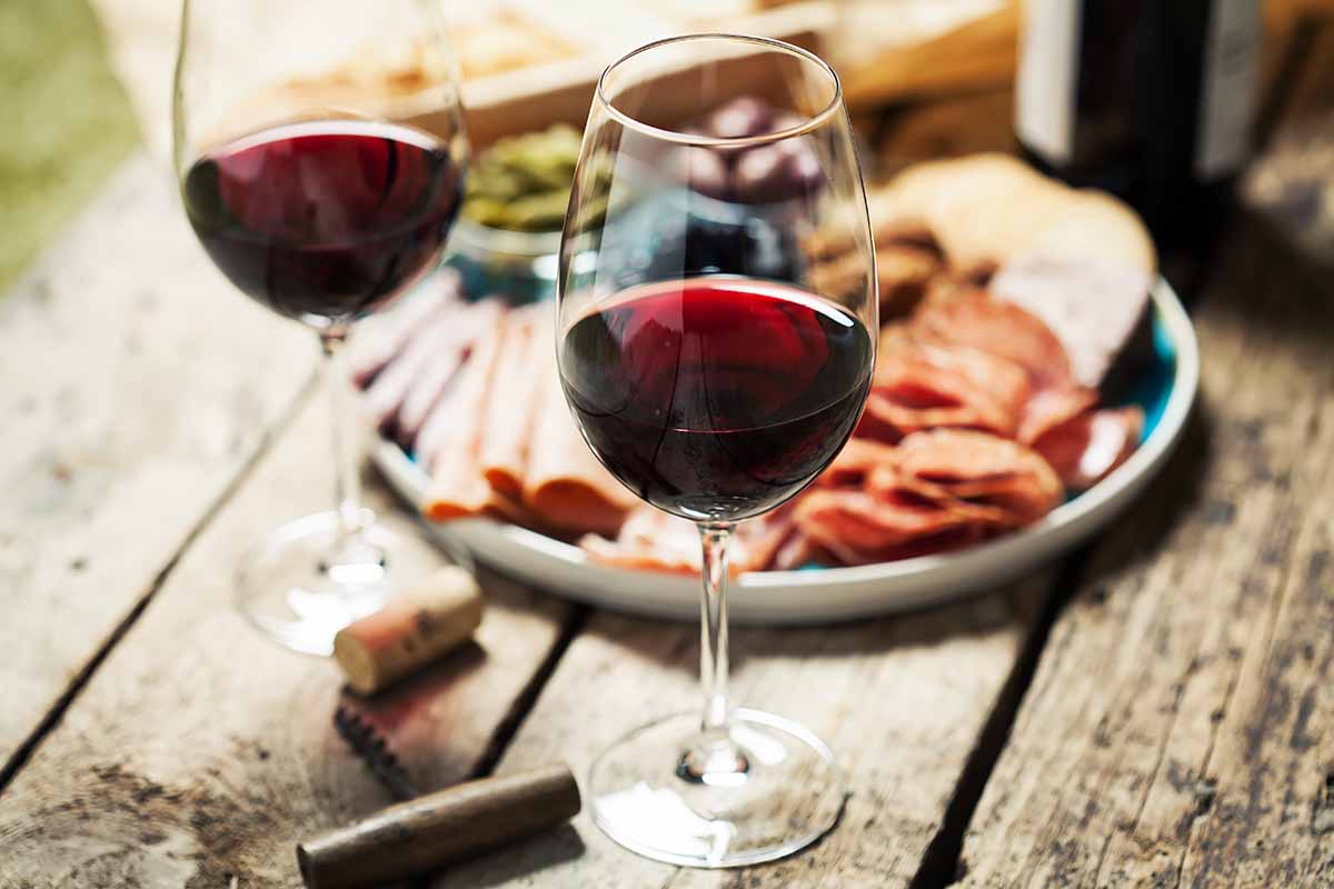 Horizontal image of two glasses filled with red wine on a dining table next to a meat and cheese plate.