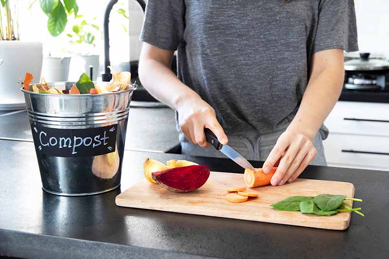 Horizontal image of preparing vegetables on a wooden cutting board next to a compost pail.