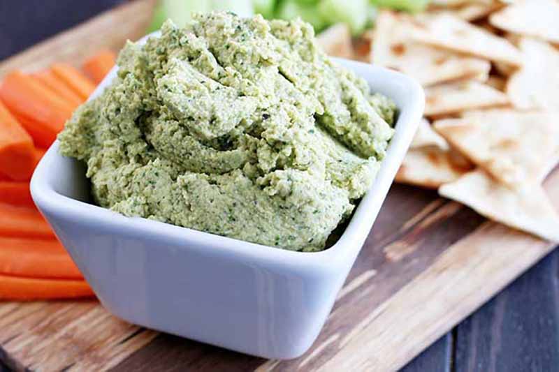 Horizontal image of a green dip in a white ramekin on a board next to carrots, celery, and pita chips.