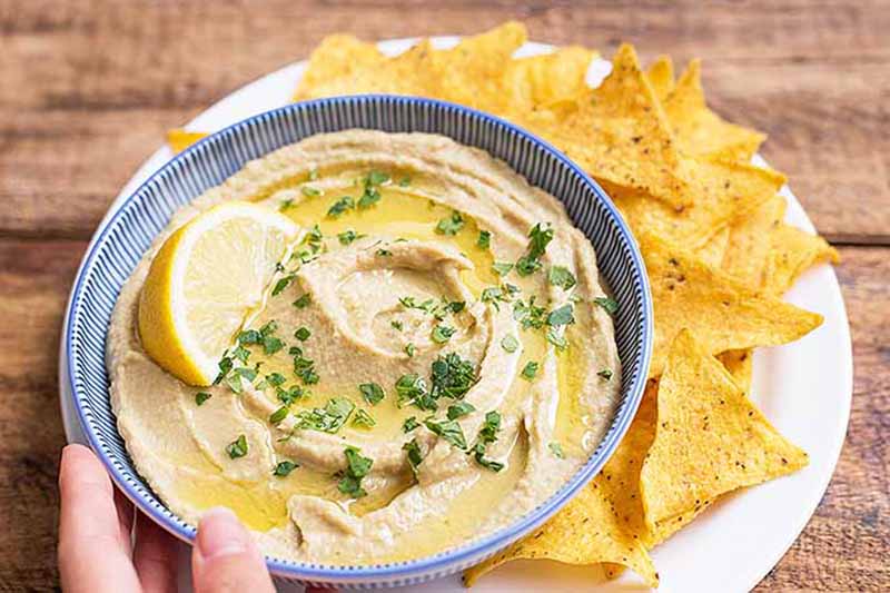 Horizontal image of a bowl filled with a hummus dip garnished with herbs and a lemon slice on a plate next to nachos.