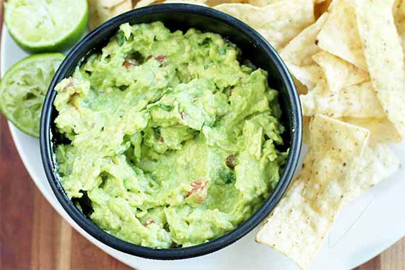 Horizontal image of a dark bowl filled with classic guacamole on a plate surrounded by nachos and limes.