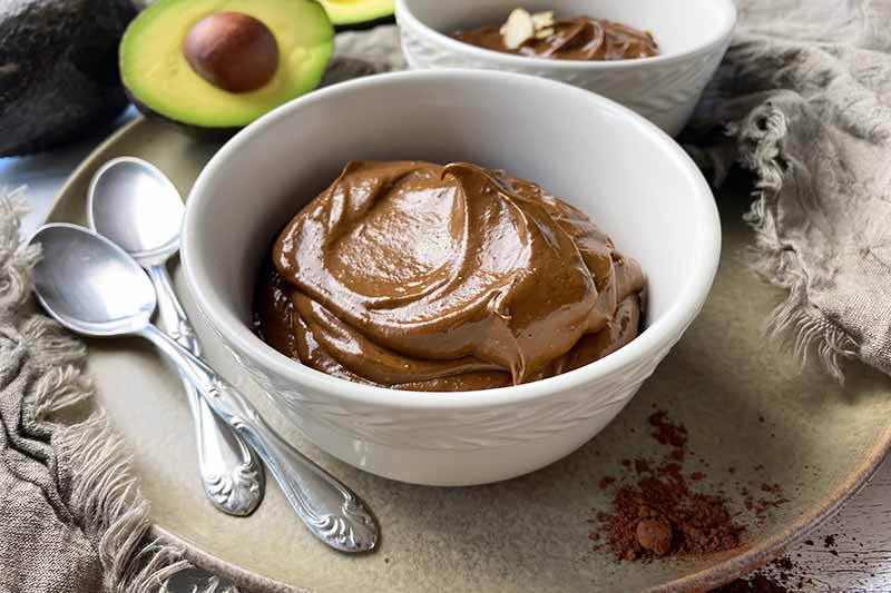 Horizontal image of two white bowls filled with a creamy cocoa dessert, one topped with sliced almonds, on a plate next to a halved avocado and spoons.
