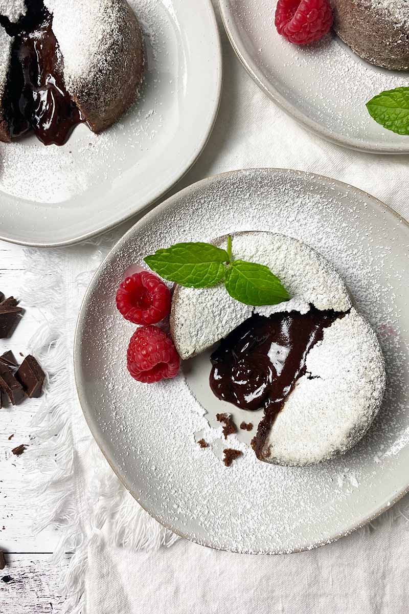 Vertical top-down image of white plates with mini cakes with a lava-like center dusted with powdered sugar and garnished with mint and raspberries.