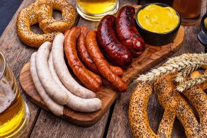 Horizontal image of three kinds of cooked meat links on a wooden board next to soft pretzels, mustard, and glasses of beer.