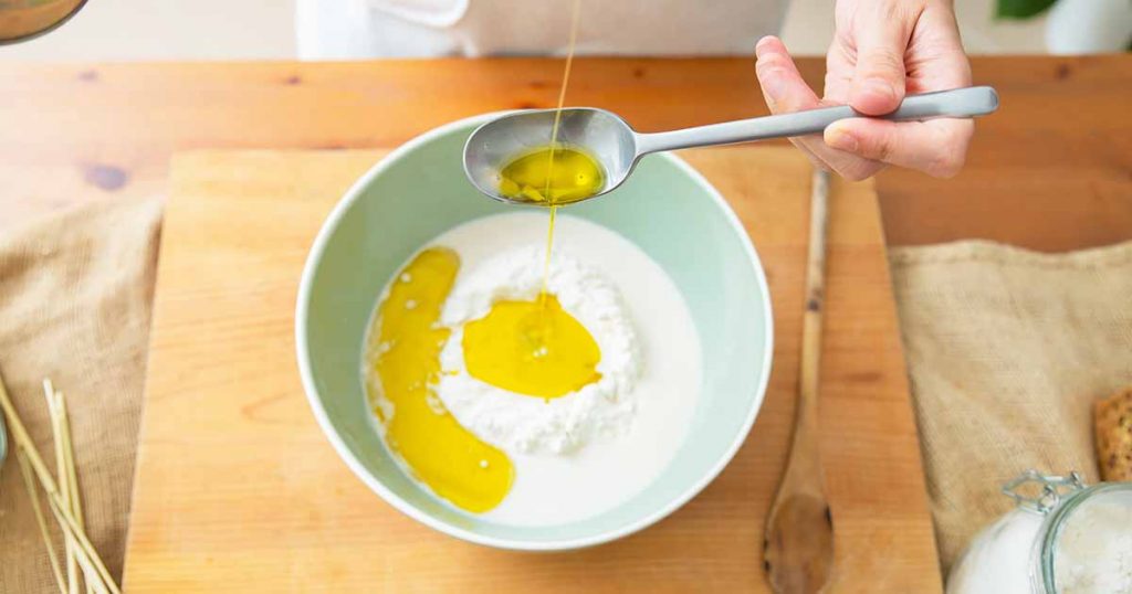 Horizontal image of pouring oil into a bowl with flour on top of a wooden cutting board.