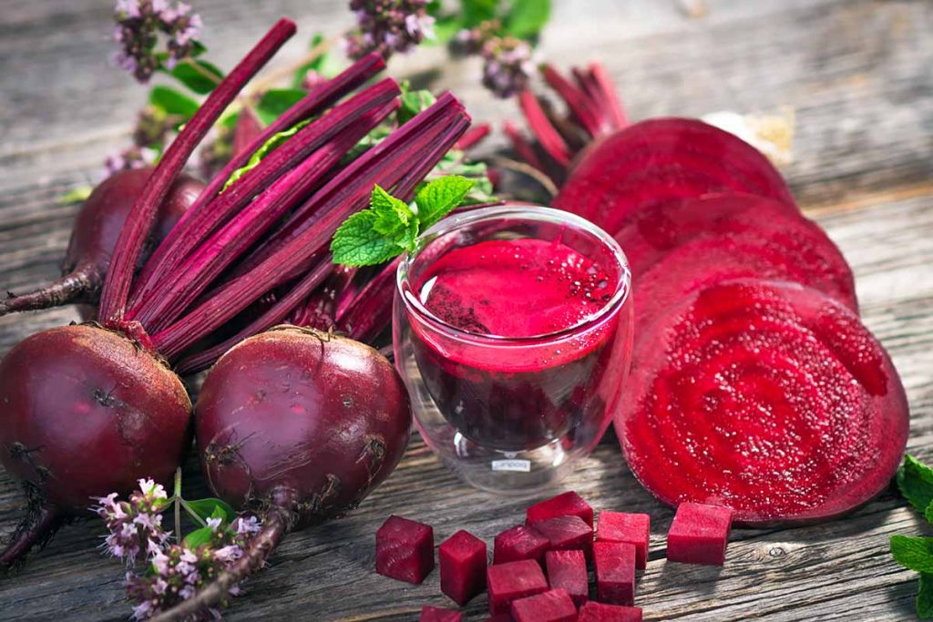Horizontal image of a glass of juice next to whole, sliced, and cubed raw red root vegetables on a wooden surface.