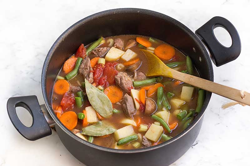 Horizontal image of cubed meat, vegetables, bay leaves, and broth in a large pot.