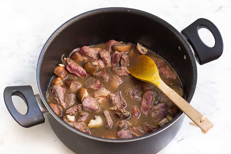 Horizontal image of cooking cubes of meat, mushrooms, and sliced onions in broth.