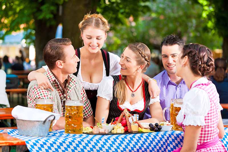 Horizontal image of a group of friends dressed in traditional German garb enjoying steins and a large charcuterie board at an outdoor table.