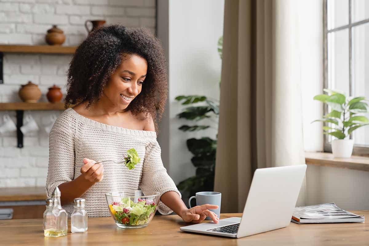 Horizontal image of a woman working on her laptop at the kitchen table eating a salad.