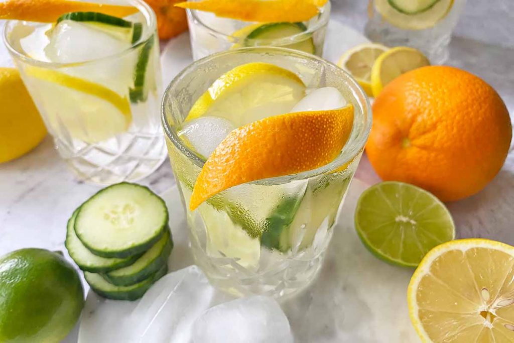 Horizontal image of glasses filled with ice, a clear liquid, and slices of fresh produce, with a thin rind as garnish.