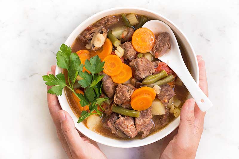 Horizontal top-down images of two hands holding the sides of a bowl filled with a hearty mix of cubed chuck and vegetables in a broth.