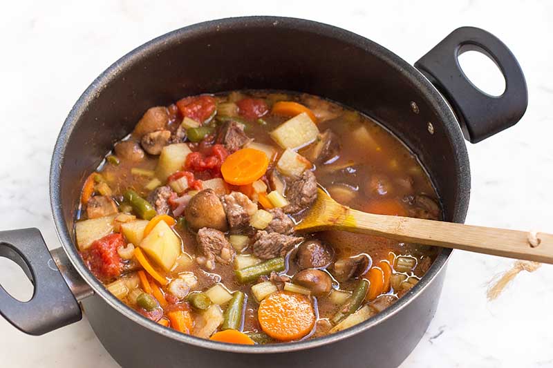 Horizontal image of a wooden spoon inserted into a large pot filled with a hearty mix of meat and veggies.