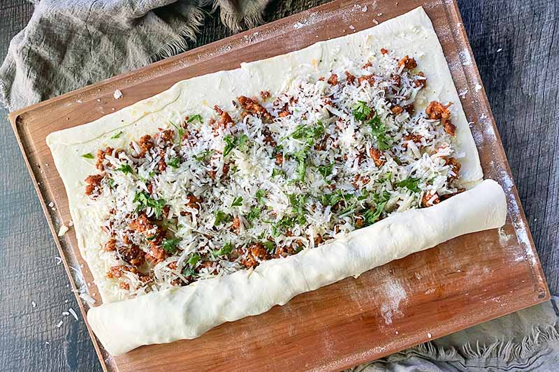 Horizontal image of rolling up a sheet of dough over a savory filling.