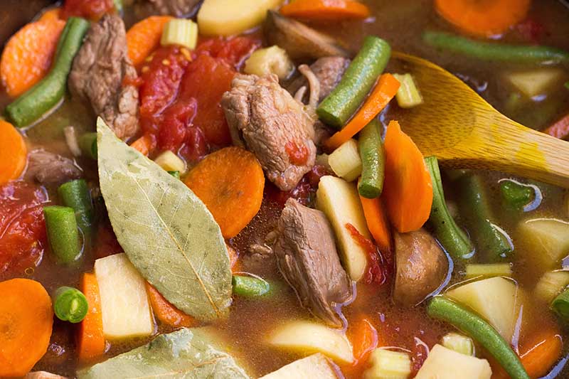 Horizontal close-up image of a hearty soup with pieces of chuck, mushrooms, carrots, green beans, potatoes, tomatoes, and bay leaves.