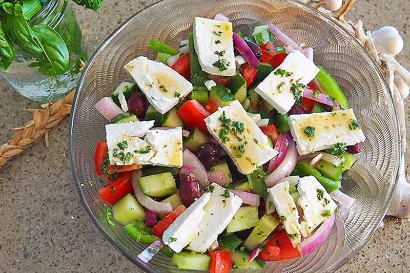 Horizontal image of a glass bowl filled with mixed vegetables topped with marinated feta cubes.