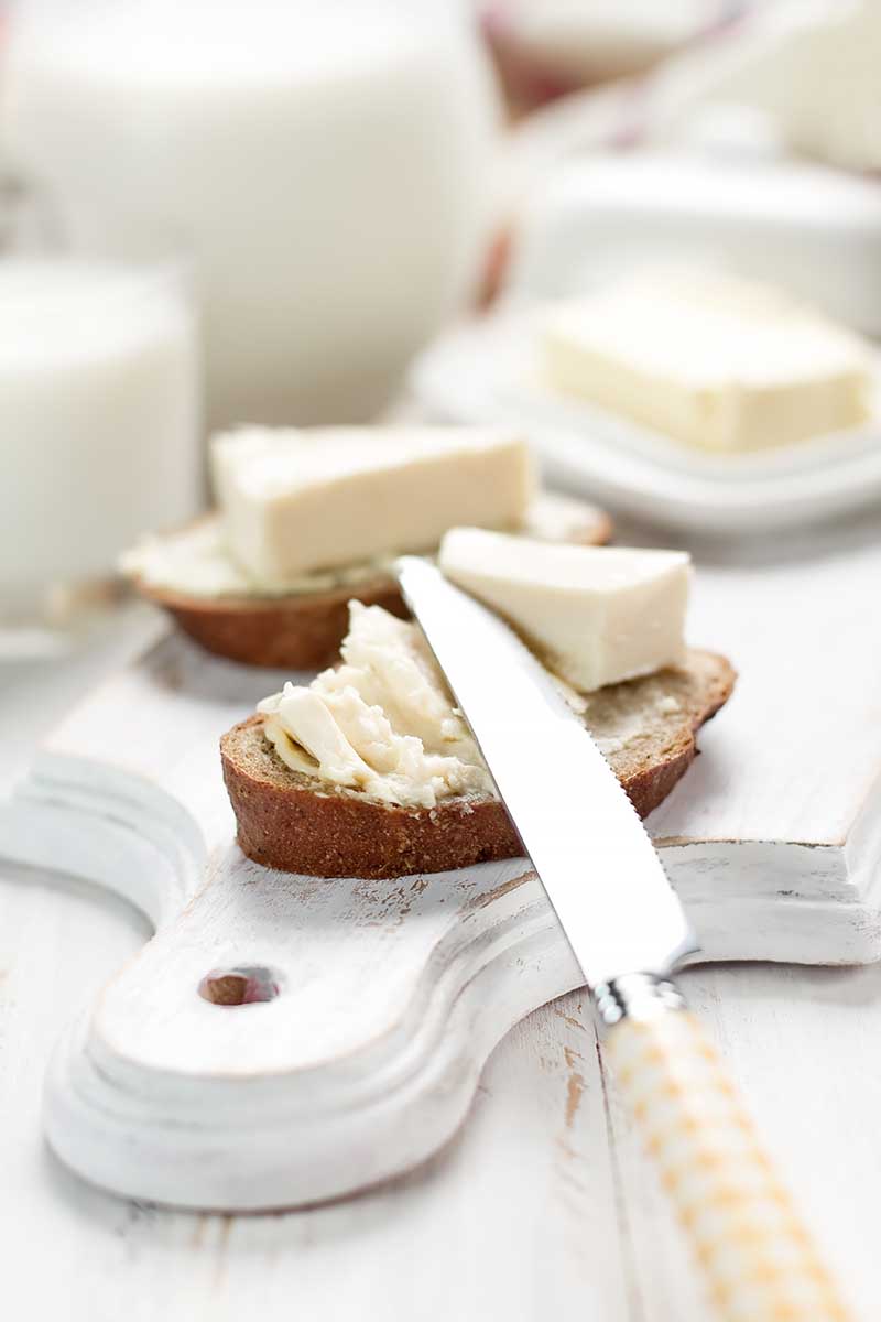 Vertical image of a knife covering slices of bread with a thick white spread on a wooden cutting board.