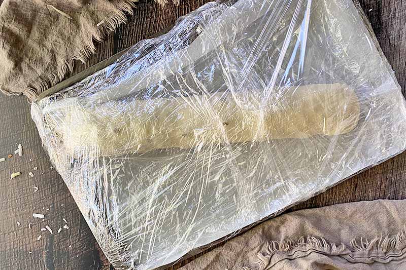 Horizontal image of an unbaked dough roll on a sheet pan wrapped with plastic wrap.