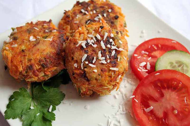 Horizontal image of three fried patties on a plate next to herbs and slices of tomatoes and cucumber.