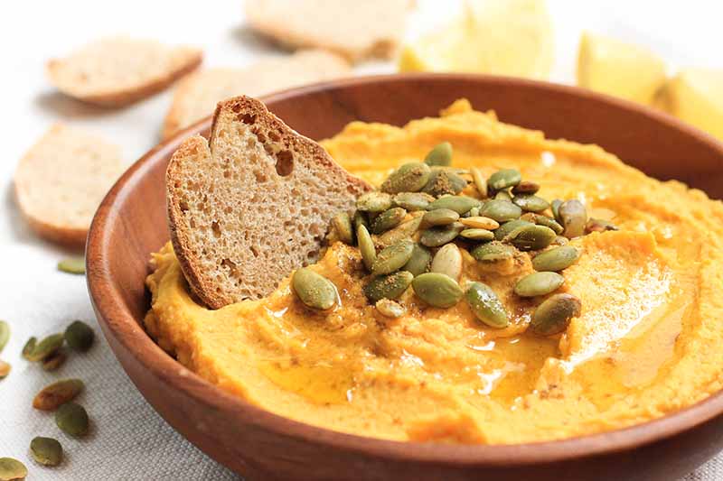 Horizontal image of a bright orange dip in a bowl topped with seeds and crackers.