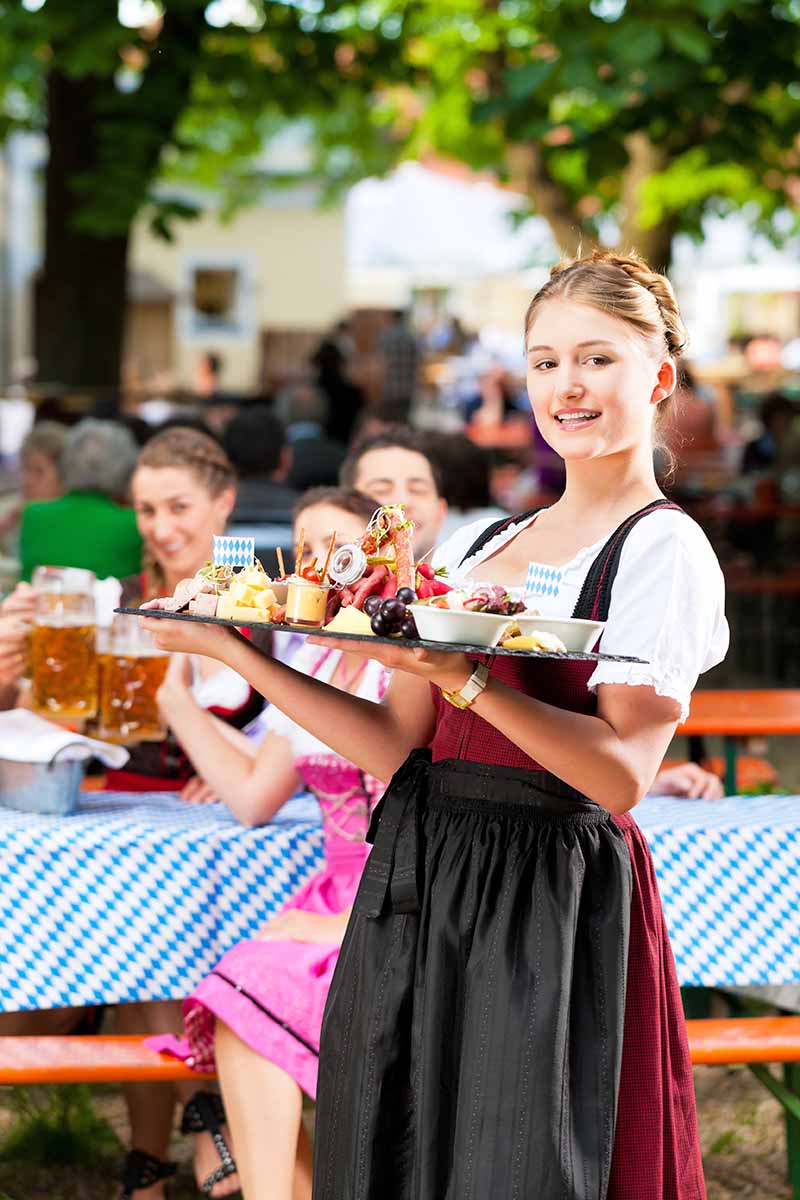 Vertical image of a female server holding a large tray of assorted charcuterie board selections in front of a table with customers drinking from large steins.