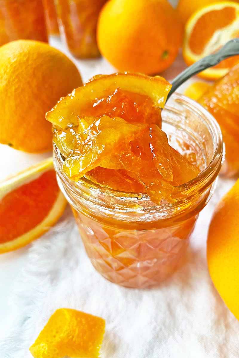 Vertical image of a spoon lifting out marmalade from a glass jar on a white towel.