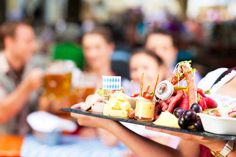 Horizontal image of a beer garden celebration with a server holding a large charcuterie board.