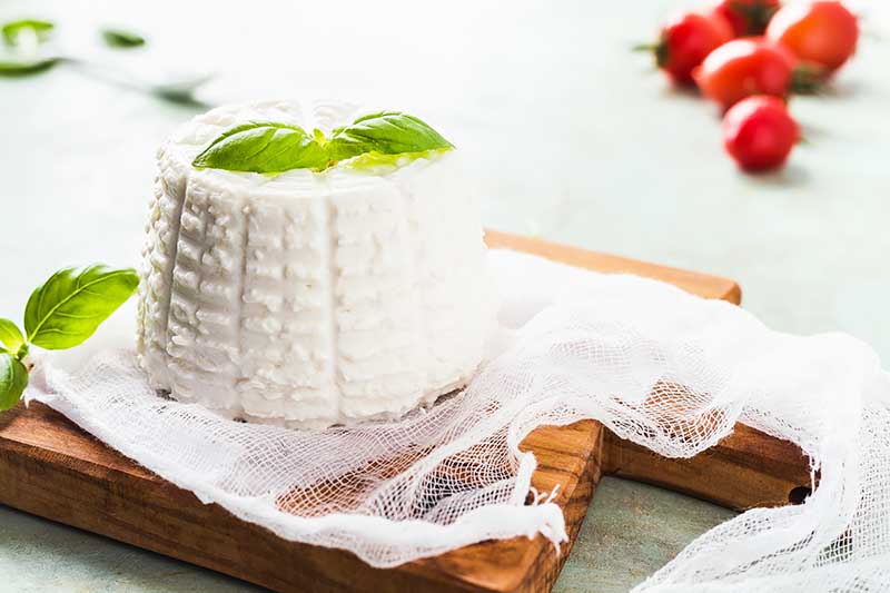Horizontal image of ricotta in the shape of a basket on cheese cloth over a wooden cutting board.