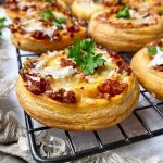 Horizontal image of rows of puff pastry rounds filled with sausage and cheese garnished with parsley on a cooling rack.