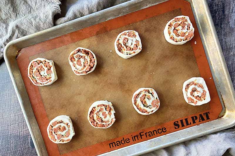 Horizontal image of two rows of unbaked dough rolled with meat crumbles on a baking sheet lined with a silicone mat.