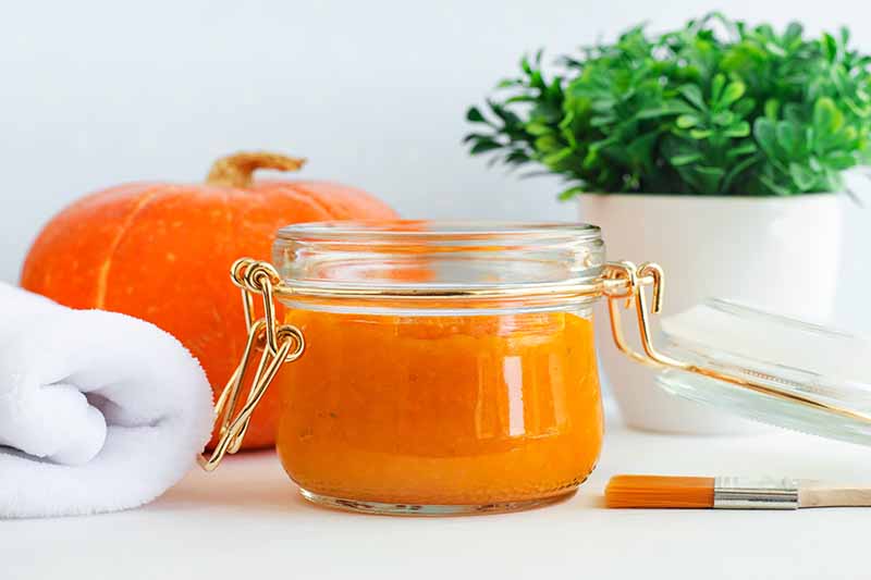 Horizontal image of a bright orange facial cream in a glass jar in front of a brush and towel.