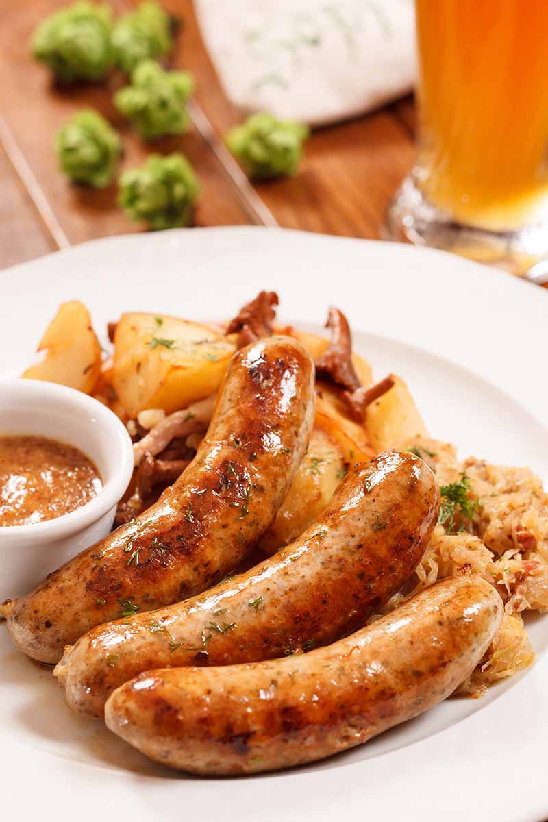 Vertical image of seared small links on a plate next to sauce and potatoes in front of a glass of beer.
