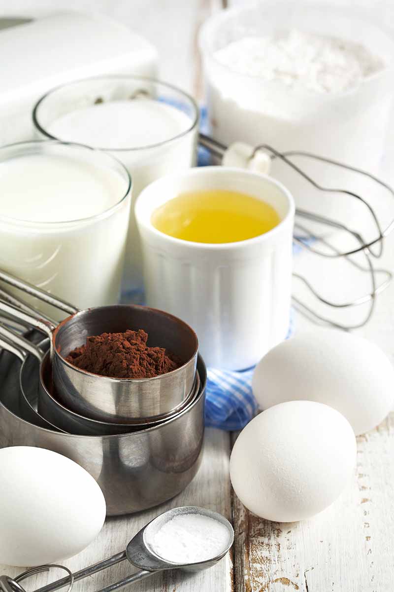 Vertical image of measured ingredients in various cups next to a whisk and eggs on a white wooden table.
