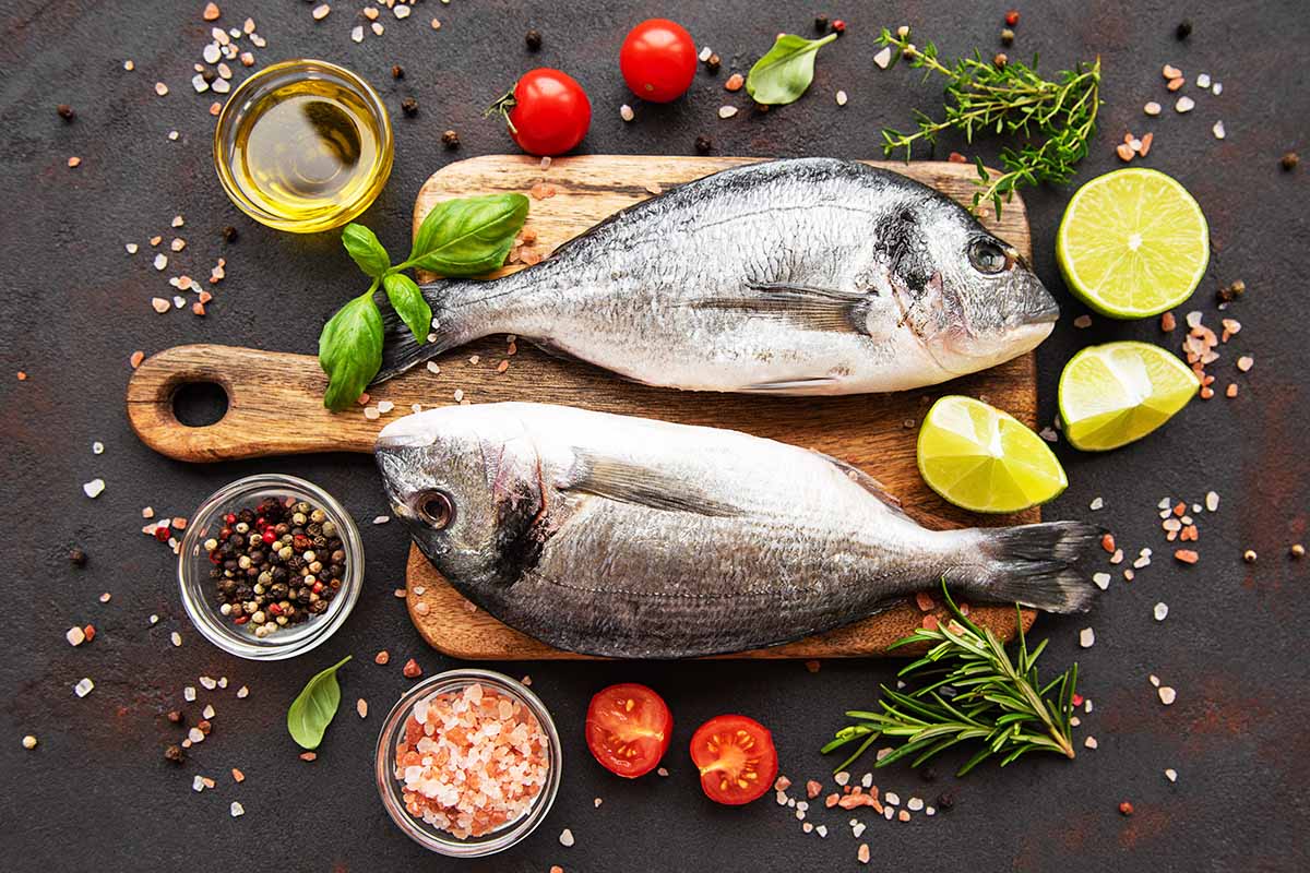 Horizontal image of two whole fish on a cutting board surrounded by tomatoes, herbs, citrus, and assorted seasonings.