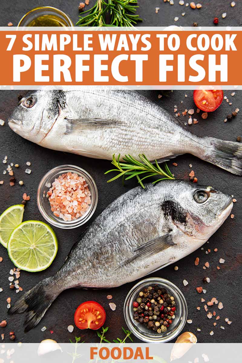 Vertical image of two whole fish on a slate surrounded by citrus, garlic, tomatoes, herbs, and assorted seasonings.