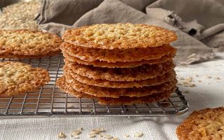Horizontal image of a stack of thin, caramelized cookies on a cooling rack over a tan napkin with oats scattered around them.