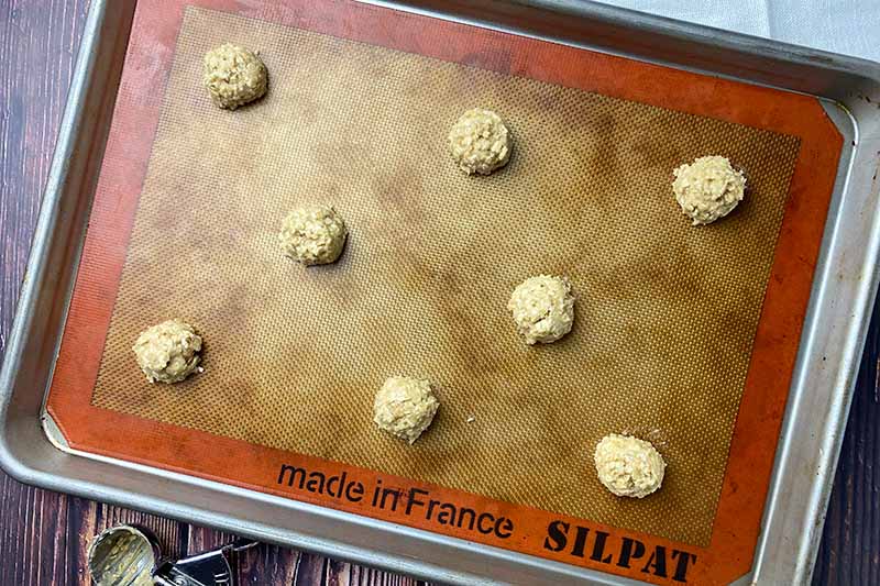 Horizontal image of unbaked rounds of batter on a baking sheet lined with a silicone mat next to a small scooper.