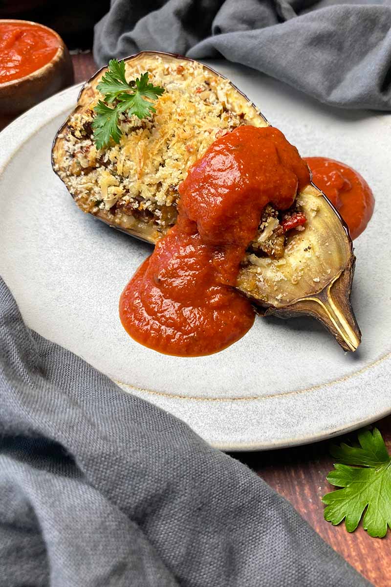 Vertical image of a stuffed eggplant half on a plate topped with tomato sauce and a parsley garnish.