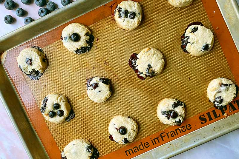 Horizontal image of baked individual cookies mixed with fresh, bursting fruit on a baking sheet lined with a silicone mat.